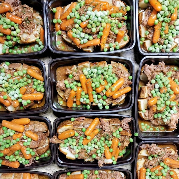 Overhead view of multiple black containers filled with pot roast, potatoes, carrots, and peas.