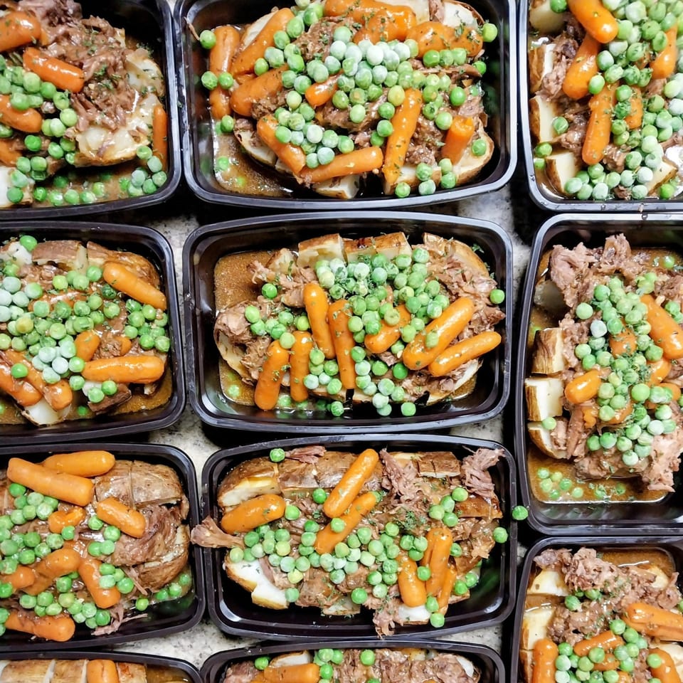 Overhead view of multiple black containers filled with pot roast, potatoes, carrots, and peas.