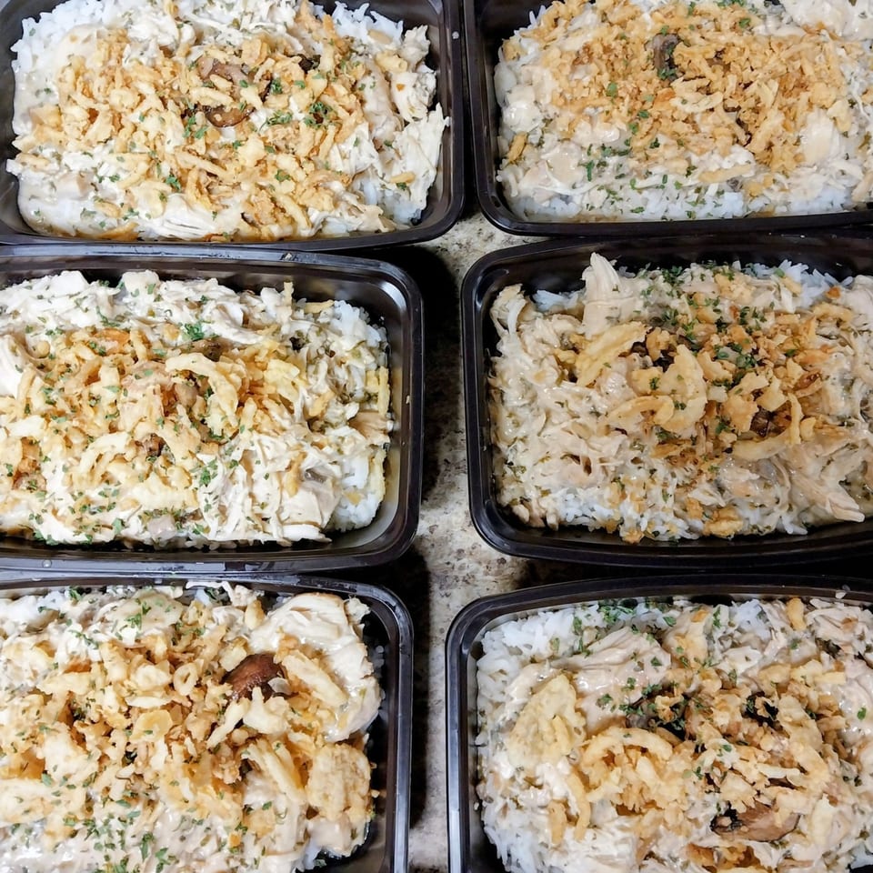 Six black containers filled with creamy chicken and rice, topped with crispy fried onions and parsley.